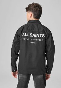 Personai ar īsiem tumšiem matiem, kas valkā melnu jaku ar baltiem uzrakstiem "ALLSAINTS" un japāņu rakstzīmēm uz muguras.