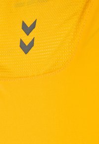 Gelbes Sportshirt mit einem strukturierten Halsbereich und drei grauen, zickzackförmigen Akzenten darunter. Glattes Gewebe ohne sichtbare Muster.