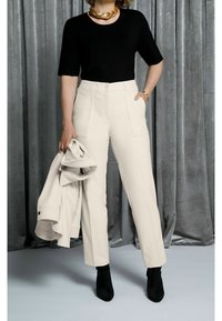 Černý krátký rukáv top v kombinaci s krémovými kalhotami na míru s předními kapsami, drženo rukou na bílé bundě. Černé kotníkové boty.