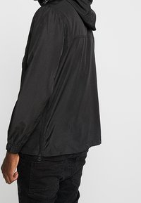 Zwarte lichtgewicht jas met capuchon, gemaakt van een glad materiaal. Voorzien van elastische manchetten en een zijzak met rits. Eenvoudig, functioneel ontwerp.