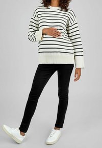 Maglione a righe bianche e nere con polsini a coste, abbinato a jeans skinny neri e sneakers bianche. La mano del modello è appoggiata sulla pancia.