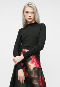 Mlada žena s dugom plavom kosom, obučena u crnu majicu dugih rukava i cvjetnu suknju s crvenim i ružičastim cvjetovima, pozira na bijeloj pozadini.