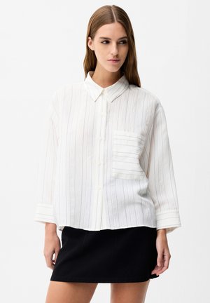 Kobieta ubrana w luźną białą koszulę zapinaną na guziki z cienkimi pionowymi paskami oraz czarną mini spódnicę, stojąca na tle jednolitego tła.