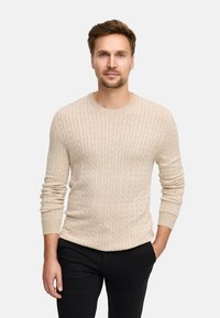 Pullover beige a maglia con maniche lunghe e scollo rotondo, caratterizzato da un motivo testurizzato e polsini a coste. Indossato con pantaloni neri.