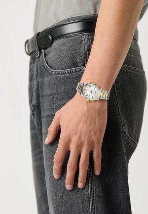 Zilveren en goudkleurige horloge met een witte wijzerplaat, Romeinse cijfers en een metalen band, weergegeven op een hand nabij zwarte jeans en een riem.