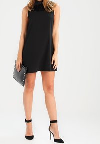 Abito nero senza maniche con un collo testurizzato, abbinato a scarpe nere con tacco alto e una pochette con borchiette. Design semplice e minimalista.