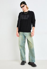 Sudadera negra con letras "GAP", vaqueros anchos de pierna en azul claro, zapatillas negras y un gorro. Conjunto informal con accesorios en capas.