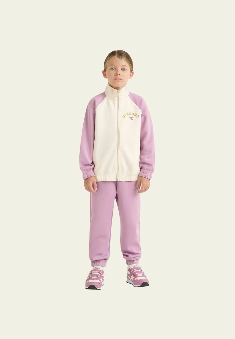 Giacca leggera con zip di colore crema chiaro, maniche viola e logo dorato, abbinata a pantaloni della tuta viola coordinati e scarpe da ginnastica rosa.