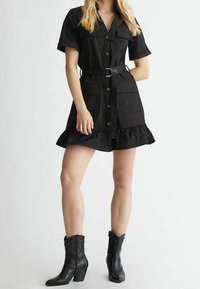 Vestito nero a camicia con maniche corte, chiusura a bottoni e due tasche. Presenta un orlo arricciato e una cintura con fibbia rotonda. Indossato con stivali neri.