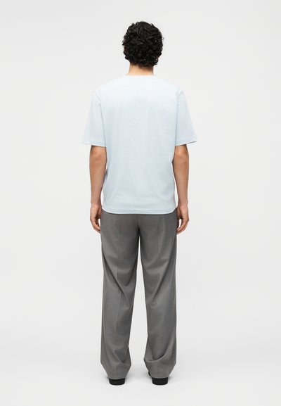 Άνδρας με σγουρά σκούρα μαλλιά που φοράει γαλάζιο κοντομάνικο πουκάμισο και γκρι φαρδιά παντελόνια, στέκεται γυρισμένος με την πλάτη σε μονόχρωμο φόντο.