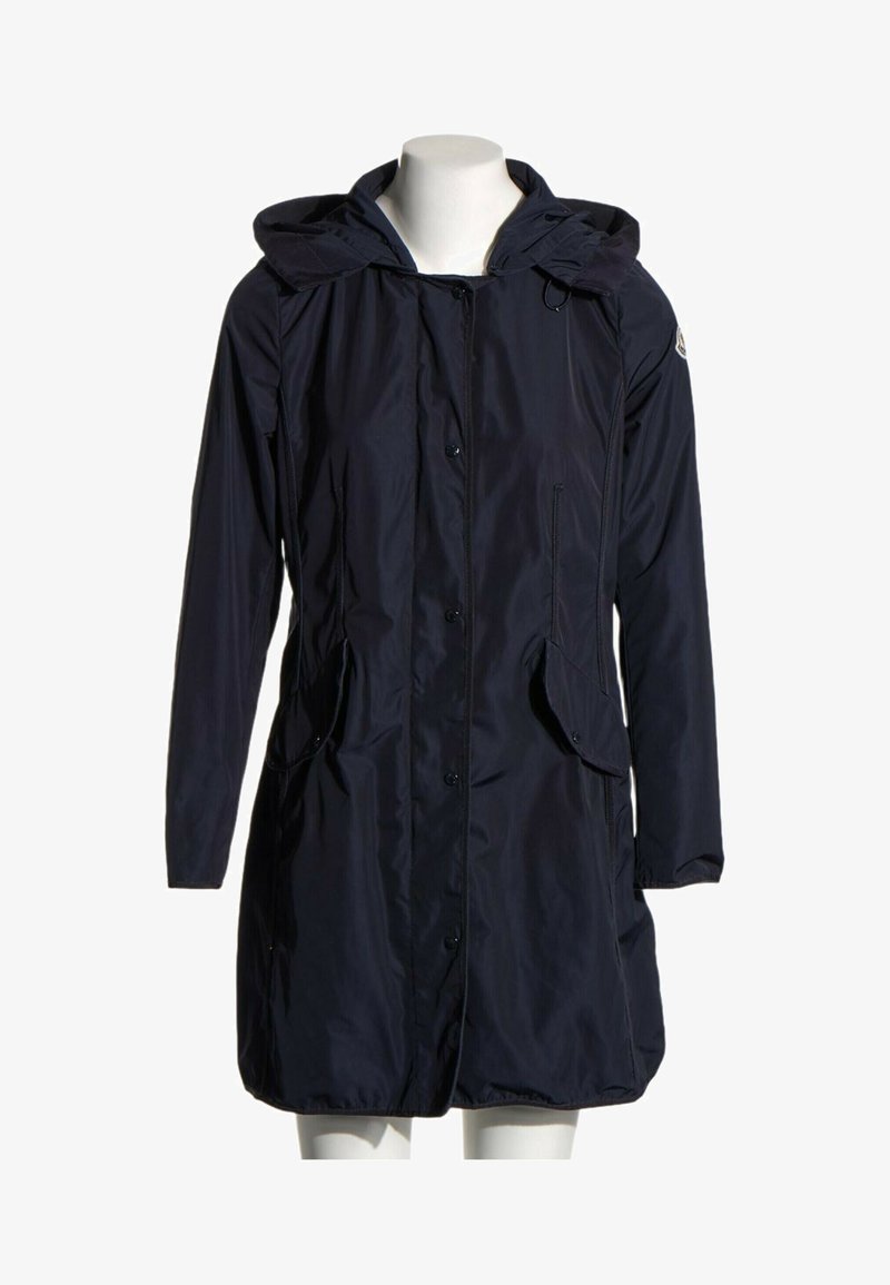 Long imperméable bleu marine foncé à capuche avec des boutons devant, des poches à rabat et un petit patch logo sur la manche gauche, présenté sur un mannequin blanc.