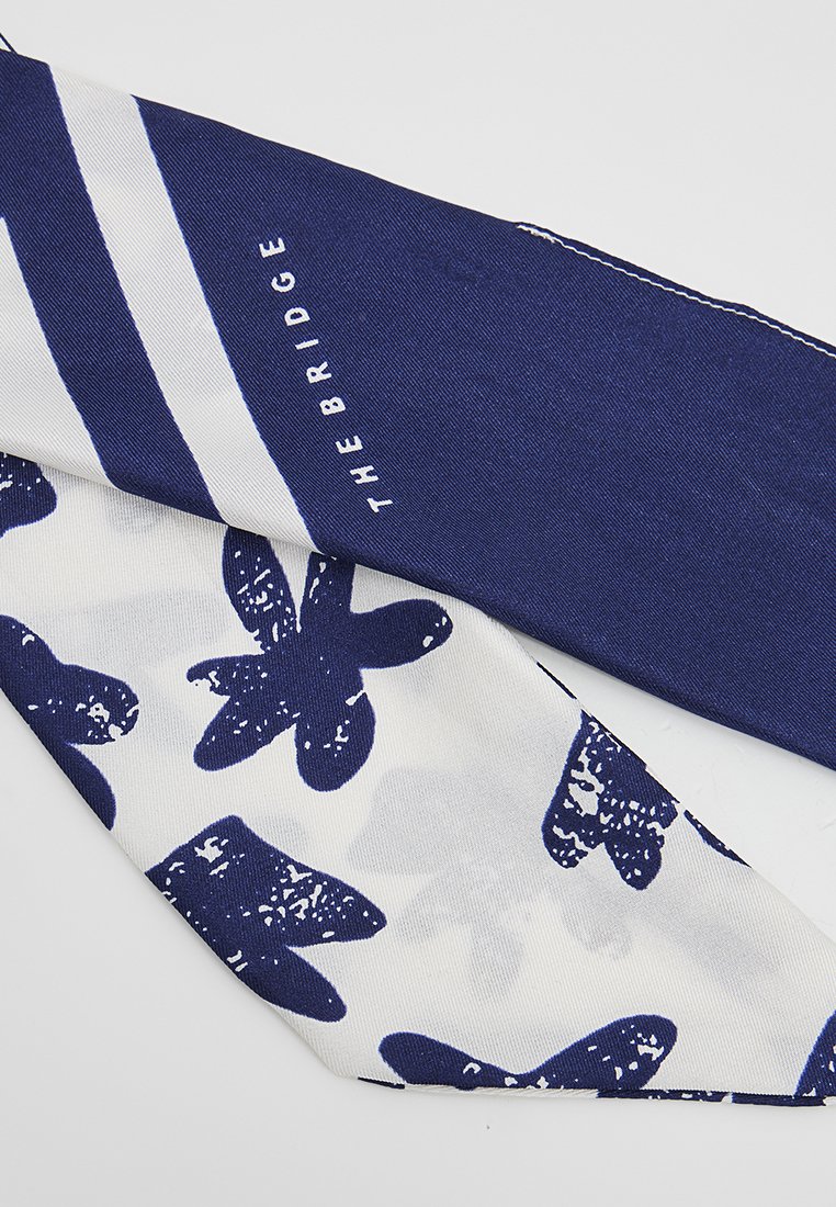 Sciarpa blu e bianca con un motivo a righe e motivi floreali. Realizzata in tessuto morbido, presenta una texture liscia e il logo 'The Bridge'.