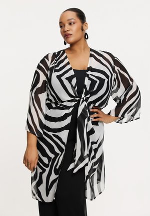 Durchsichtiger Kimono im schwarz-weißen Zebramuster, mit weitem Ärmel, gebundenem Vorderteil und fließender Silhouette. Kombiniert mit einem schwarzen Top.