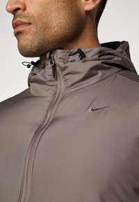 Nike-jacka i ljusgrått tyg med blixtlås, huva med justerbara snoddar och en liten broderad logga på bröstet.