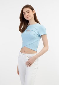 Světle modrý žebrovaný crop top s krátkými rukávy, se asymetrickým střihem a předním výřezem. Kombinované s bílými kalhotami.