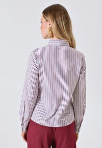 Camicia a maniche lunghe bianca con righe verticali bordeaux, dotata di colletto e polsini con bottoni, abbinata a pantaloni borgogna.