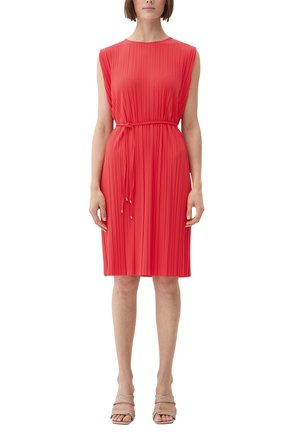 Kobieta w bezrękawowej, koralowej, plisowanej sukience do kolan z cienkim paskiem i beżowych sandałach na otwartym palcu, stoi na tle białym.