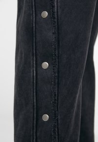 Sorte denimbukser med rå kant og sølvtryklåse langs siden, der fremhæver en glat tekstur og et strømlinet design.