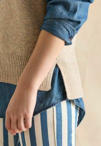 Beige Strickpullover über einem blauen Jeanshemd, mit gestreiften weißen und blauen Hosen. Merkmale sind ein gerippter Saum und seitliche Schlitze für zusätzliche Details.