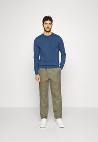 Sweater azul de manga longa com decote redondo, combinado com calças de tom oliva claro e ténis brancos, criando um visual casual.