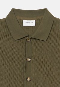 A camisa polo em verde oliva, tricotada, apresenta um padrão texturizado, um colarinho canelado e três botões castanhos na parte frontal. Etiqueta: Les Deux.