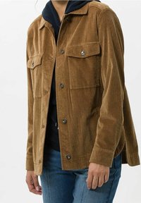 Giacca in velluto a coste marrone con due tasche sul petto, bottoni a pressione argentati e una vestibilità comoda. Indossata sopra un felpa scura e jeans.