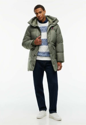 Giovane uomo con giacca imbottita verde oliva sopra un maglione bianco e blu a fantasia, jeans scuri e sneakers bianche, in piedi e in posa.