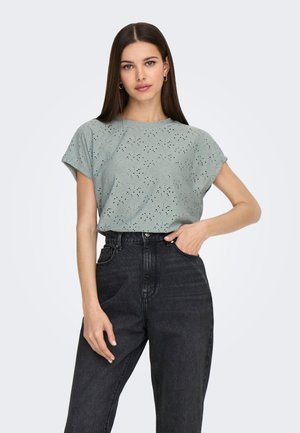 Kobieta o długich brązowych włosach, ubrana w jasnorozzielony ażurowy top z krótkim rękawem i wysokim stanem czarne jeansy, z jedną ręką w kieszeni.