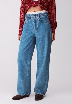 Persona che indossa jeans blu a vita alta e gamba larga abbinati a scarpe a punta marrone scuro e una maglia a maniche lunghe rossa con motivi, annodata in vita.