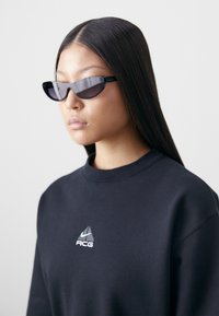 Black sunglasses with a narrow, angular design and tinted lenses. Model wears a black sweatshirt featuring a logo on the chest.