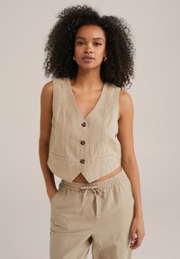 Beige lněná zkrácená vesta s V-šedým výstřihem, se třemi tmavými knoflíky a bočními kapsami, doplněná volným střihem.