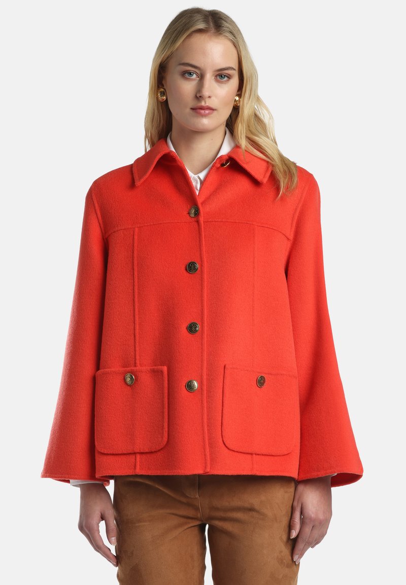 Raudonas vilnonis pelerinos paltas su apykakle, auksiniais mygtukais ir dviem priekinėmis kišenėmis. Turi boxy siluetą ir lygų paviršių.