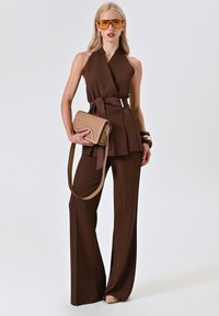 Braunes Halsbandoberteil mit gebundenem Bund, kombiniert mit weitem Beinkleid. Accessoires sind eine geflochtene Clutch und übergroße gelbe Sonnenbrillen.