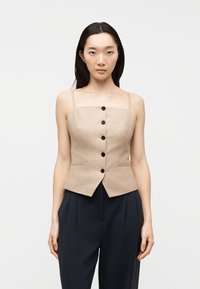 Femme portant un haut beige à rayures fines sans manches boutonné et un pantalon noir taille haute, debout devant un fond clair uni.