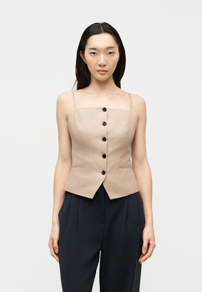 Femme portant un haut beige à rayures fines sans manches boutonné et un pantalon noir taille haute, debout devant un fond clair uni.