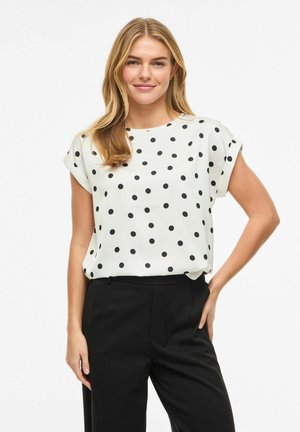 Młoda kobieta o blond włosach, ubrana w białą bluzkę z krótkim rękawem w czarne grochy oraz czarne spodnie, stoi z jedną ręką na biodrze.