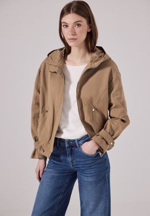Veste kaki avec capuche, dotée d'une fermeture éclair à l'avant et de boutons-pression, associée à un haut blanc et à un jean large bleu. Tissu lisse, coupe décontractée.