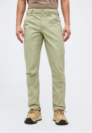 Muškarac obučen u svijetlozelene hlače ravnog kroja i smeđe planinarske cipele, stoji ispred jednobojne svijetlosive pozadine.