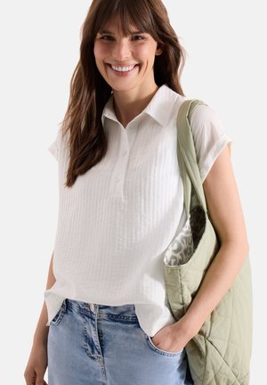 Femme souriante portant un chemisier blanc à manches courtes et un jean bleu, une main dans la poche, portant un sac cabas matelassé vert clair sur l'épaule.