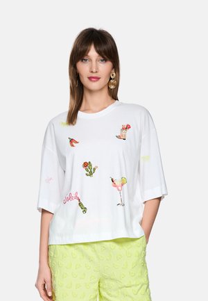 Frau trägt weißes übergroßes T-Shirt mit bunten bestickten Mustern und hellgrünen, strukturierten Shorts, steht mit einer Hand in der Tasche.