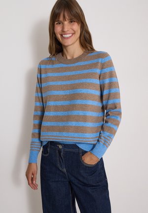 Femme souriante portant un pull à rayures marron et bleu à manches longues et un jean bleu foncé, une main dans la poche, sur un fond uni.
