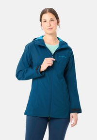 Femme portant une veste à capuche bleue, tenant la fermeture éclair près de la poitrine, debout devant un fond clair uni.