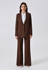 Blazer marrone con un solo bottone e rever a punta, abbinato a pantaloni larghi coordinati. Camicia bianca con colletto sotto. Tessuto liscio.