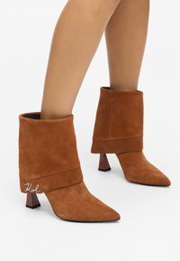 Stivaletti ankle in suede marrone con risvolto, punta affusolata e tacco a blocco trasparente; presentano un dettaglio del logo argentato sul risvolto.