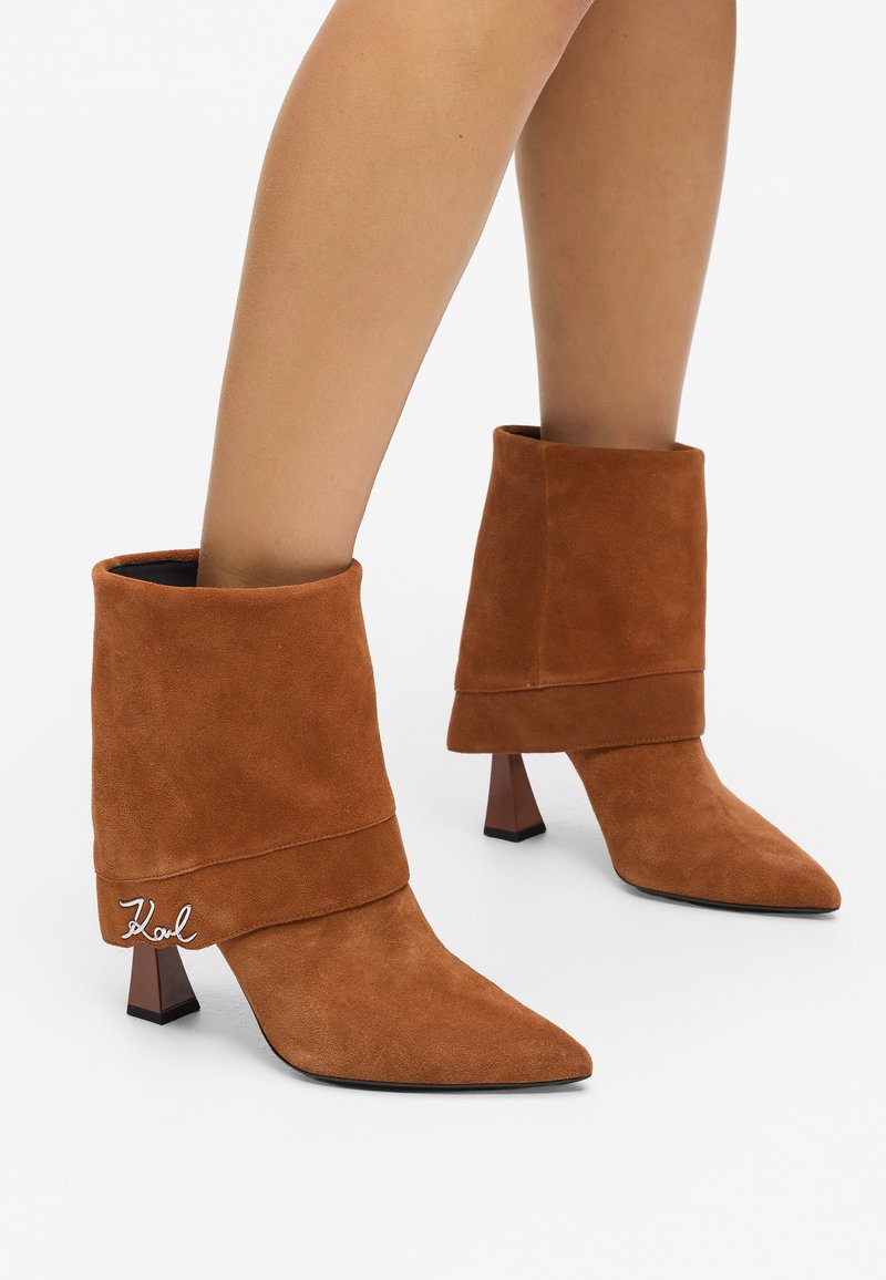 Stivaletti ankle in suede marrone con risvolto, punta affusolata e tacco a blocco trasparente; presentano un dettaglio del logo argentato sul risvolto.