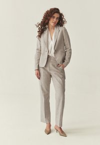 Mujer con cabello rizado que lleva un traje gris claro con pantalones a medida, una blusa blanca y tacones beige puntiagudos, de pie contra un fondo liso.