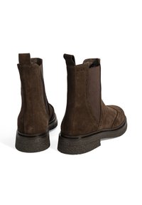 Bottines en daim marron avec des panneaux élastiques sur les côtés, semelle en caoutchouc texturé et détails perforés à l'avant. Vue arrière montrée.