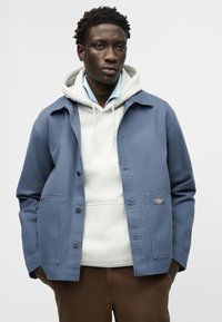 Νεαρός άνδρας που φοράει ανοιχτό μπλε μπουφάν με κουμπιά πάνω από λευκή κουκούλα και καφέ παντελόνι, μπροστά σε απλό γκρι φόντο.