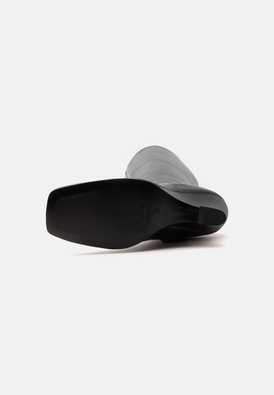 Botte en cuir noire vue de la semelle avec un bout carré et un petit talon bloc sur un fond blanc uni.