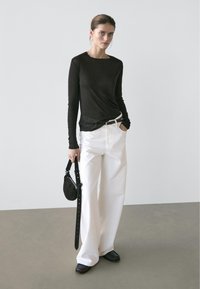 Woman standing against a plain wall wearing a black long-sleeve top, white wide-leg pants, black shoes, holding a small black handbag.